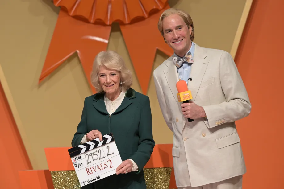The Queen smiles alongside actor Oliver Chris whilst holding a clapper board